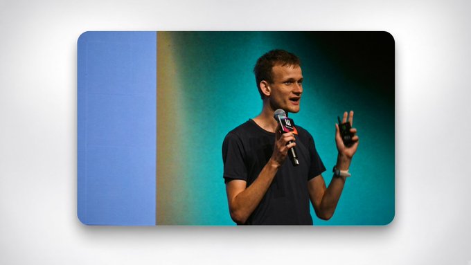 A man wearing a black t-shirt stands on a stage holding a microphone and a clicker, presenting against a blue and green backdrop.