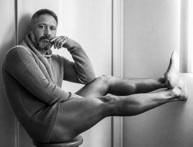 A black-and-white portrait of a bearded man in a knit sweater posing with his legs extended against a wall.