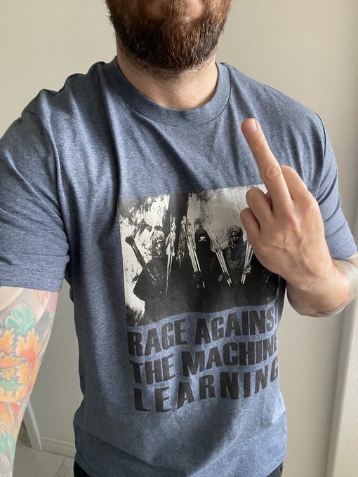 A bearded man wearing a blue t-shirt that reads 'RAGE AGAINST THE MACHINE LEARNING' with a band photo, while pointing his finger upward.