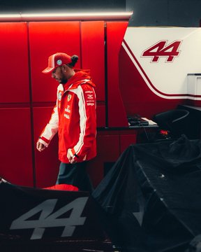 Lewis Hamilton, wearing his red Ferrari cap and team jacket, walks through the paddock as the number 44 car is visible, gearing up for another race day with Scuderia Ferrari.