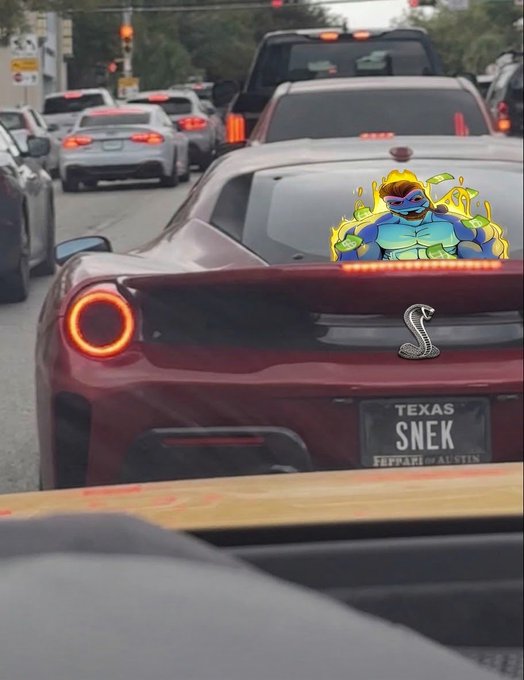 A red Ferrari sports car with a Texas license plate reading 'SNEK' being driven in traffic, with an edited cartoon character graphic overlaid on the back window.