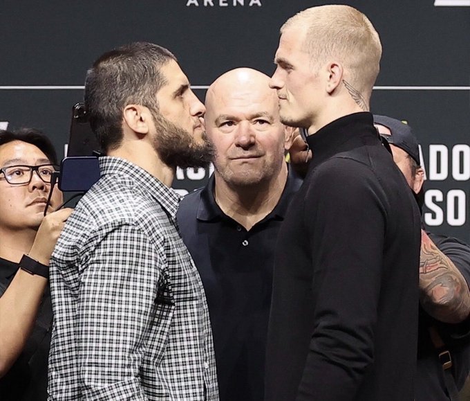 Two MMA fighters face off in a staredown at a press event, with Dana White standing between them. The text 'ARENA' is partially visible in the background.