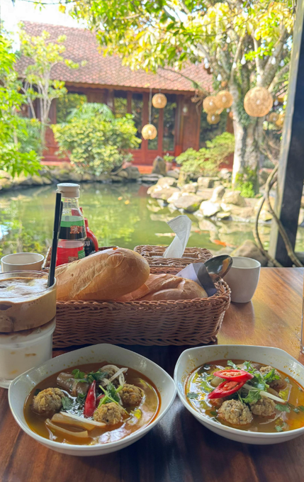 A table set with two bowls of meatball soup, bread in a basket, and drinks, overlooking a pond and garden.