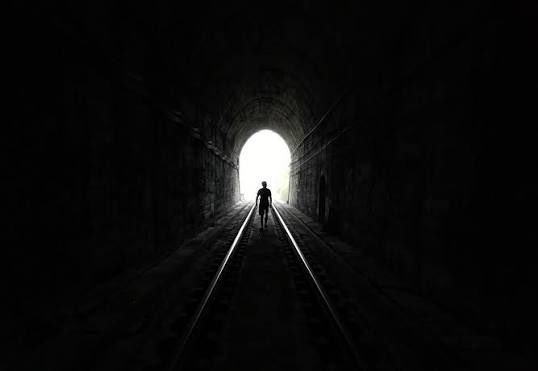 A dark, atmospheric silhouette of a person standing on train tracks inside a tunnel, with a bright opening at the far end.