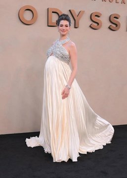 The image shows a pregnant woman posing on the red carpet at the "ODYSSEY" premiere, wearing an elegant white Grecian-style gown with a beaded bodice that highlights her baby bump. Anne Hathaway announced her third pregnancy on June 19, with gender prediction markets on polymarket.com showing slightly favoring a boy, making this red carpet appearance a notable public reveal of her pregnancy.