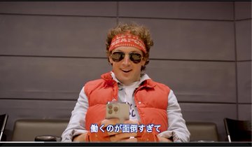 A video still of a man wearing a red bandana, sunglasses, and red vest sitting at a desk holding a plush toy, with Japanese subtitle text 驚くのが面倒すぎて.