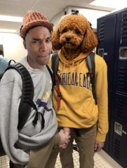 Two men pose in a school hallway, one wearing a beanie and gray sweatshirt, the other in a yellow 'American Eagle' hoodie with a large brown teddy bear mask over his head.