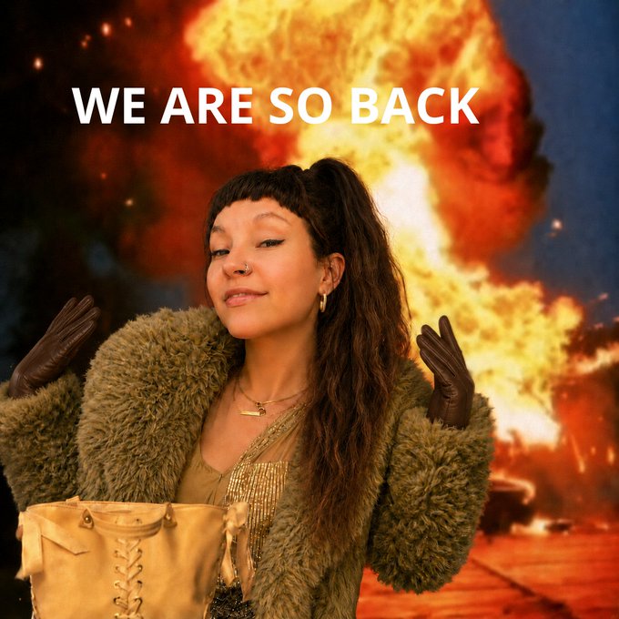 A woman with curly dark hair and bangs poses confidently in front of a large explosion backdrop, wearing a fuzzy green coat and brown gloves, with the bold white text 'WE ARE SO BACK' displayed above her.