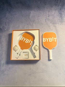 A custom orange pickleball paddle branded with the "BYBIT" logo rests beside its matching orange packaging on a gray surface.