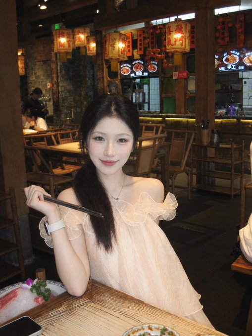 A young woman with long dark hair wearing a sheer, off-the-shoulder top holds chopsticks while dining at a restaurant decorated with traditional Chinese lanterns.
