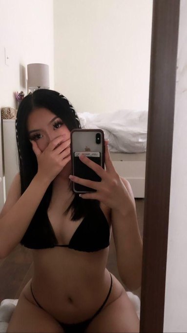 A woman in black lingerie taking a mirror selfie with a smartphone in a bedroom.