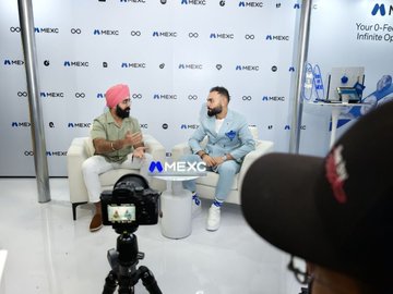 The image captures a candid interview between two men seated in white chairs in front of a backdrop and table branded with the "MEXC" logo, with a camera recording the conversation. The setup reflects the post's reference to a podcast recording with "usithetalk," featuring Vugar sharing insights for MEXC.