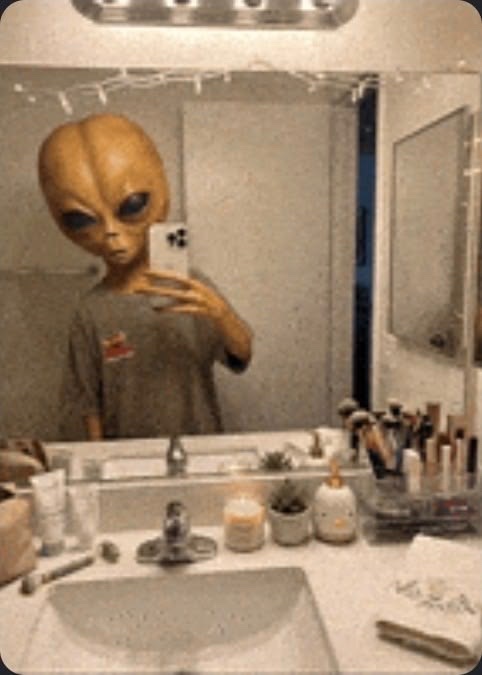 A person in an alien mask taking a mirror selfie in a bathroom with a phone, wearing a gray shirt, with various toiletries on the counter.