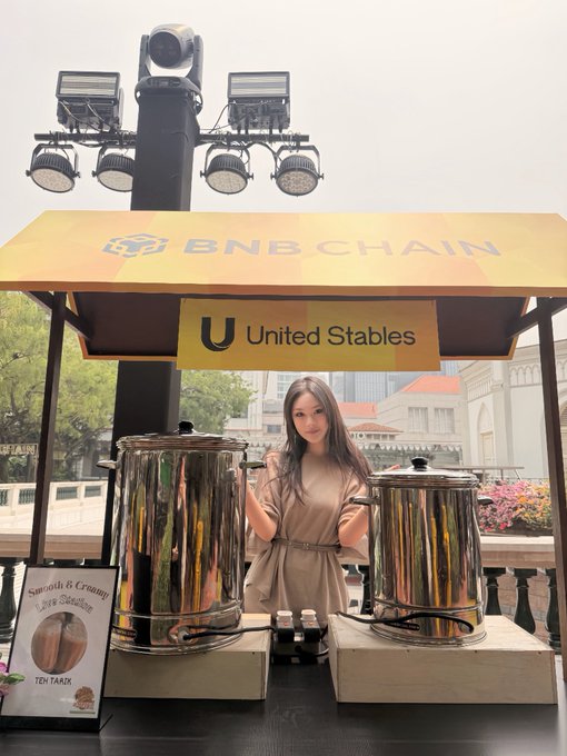 A woman poses behind a milk tea station sponsored by BNB Chain and United Stables, ready to serve creamy "Teh Tarik" as part of the BNB Singapore event. The hashtag #BNBSG highlights the local promotion, blending blockchain branding with a playful, welcoming drink experience.