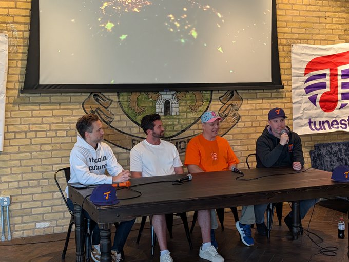 Four men sit at a table during a panel discussion in a room with brick walls, a projector screen, and a "tunesi..." banner visible in the background.