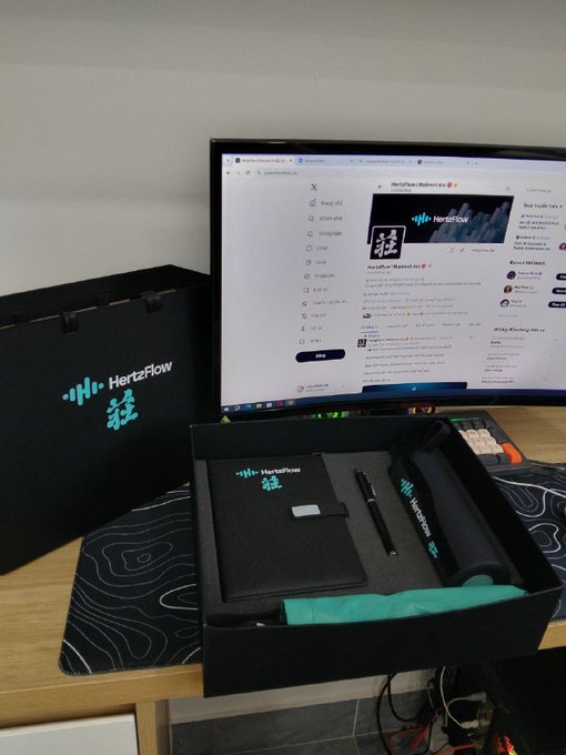 Photo of a 'HertzFlow' branded welcome kit on a desk, featuring a black gift bag, a notebook, pen, water bottle, and a green item inside a black presentation box, with a monitor displaying a Twitter profile in the background.