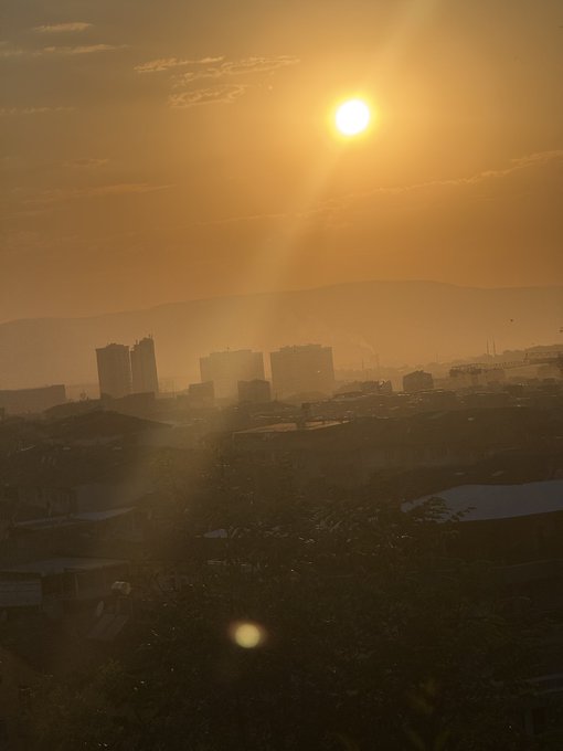 A golden sunrise casts a warm glow over a city skyline, with the sun's rays creating a beautiful haze and lens flare. A perfect way to say "Gm fam" and greet the new day.