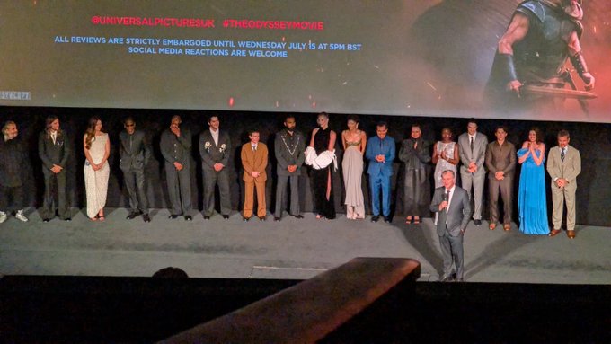 Cast of 'The Odyssey' standing on stage in front of a screen showing review embargo information from Universal Pictures UK, with the text 'ALL REVIEWS ARE STRICTLY EMBARGOED UNTIL WEDNESDAY JULY 15 AT 5PM BST'.