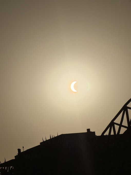 A partial solar eclipse glows brightly in a hazy sky above the silhouette of a metal bridge, likely captured with a camera pointed directly at the sun—a likely reason the camera no longer works.