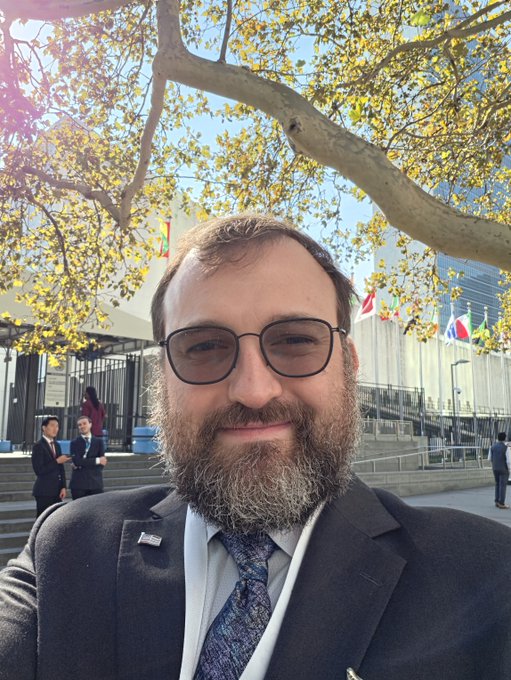 A bearded man in a dark suit, sunglasses, and a blue patterned tie takes a selfie in front of the United Nations headquarters, where member state flags can be seen in the background near the iconic building. The image, captioned "Good to be here at the United Nations," reflects a professional visit to the UN headquarters.
