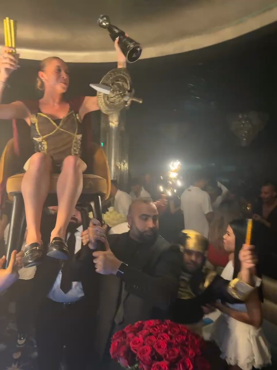 A woman in a dark dress sits on an ornate chair being lifted by men, holding a bottle aloft, while someone holds up a sparkler and a large bouquet of red roses sits below.