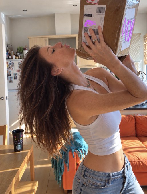 A woman with long brown hair wearing a white tank top and jeans drinks from a large cardboard box in a sunlit living room.