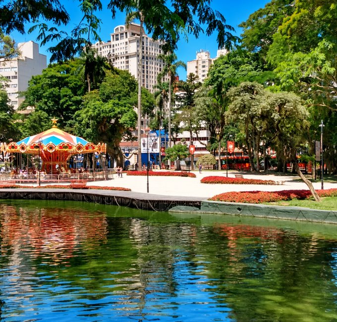 A scenic park view featuring a colorful carousel, lush green trees, a reflective pond in the foreground, and city buildings visible in the background.