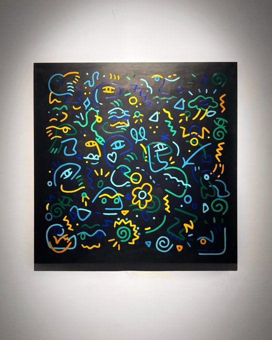 The image features a large abstract painting with various shapes and colors, including blue, green, yellow, and orange lines. The artwork is displayed prominently against a white wall, making it stand out as the main focus of the room. The artist's signature can be seen in the bottom right corner of the painting.