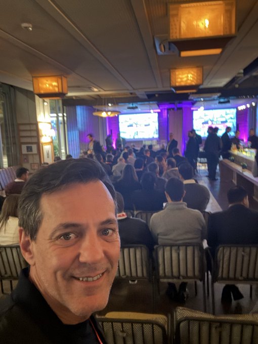 A man takes a selfie at what appears to be a conference or event, with a crowd of seated attendees and presentation screens visible in the background.