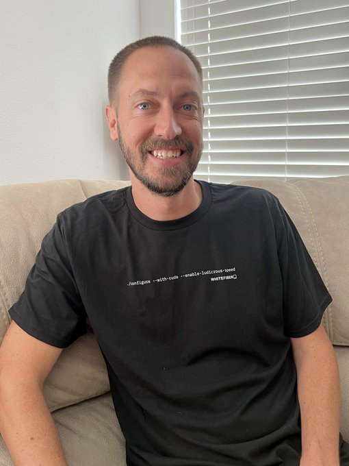 A smiling man with a short beard wearing a black t-shirt with small white command-line text sits on a beige couch.