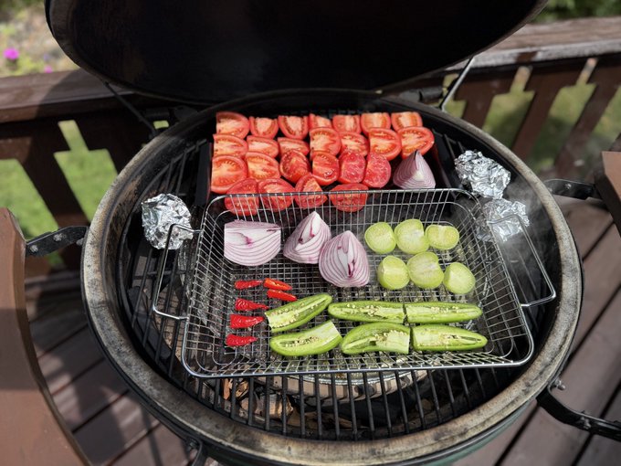 A kamado-style grill with a wire mesh basket containing sliced tomatoes, onions, cucumbers, jalapeños, and foil-wrapped items cooking over hot coals.