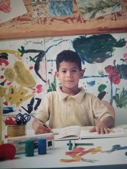 A childhood photograph of a young boy smiling while sitting at a desk with an open notebook, colorful paints, and scissors, mounted on a corkboard surrounded by children's artwork.