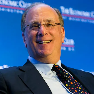 A portrait photograph of a smiling man wearing glasses, a dark suit, and a polka dot tie, photographed at 'The Economic Club' event with a blue background.