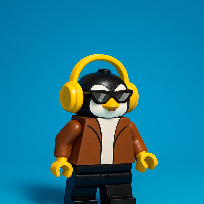 A LEGO-style penguin figure wearing yellow headphones, sunglasses, and a brown jacket against a solid blue background.