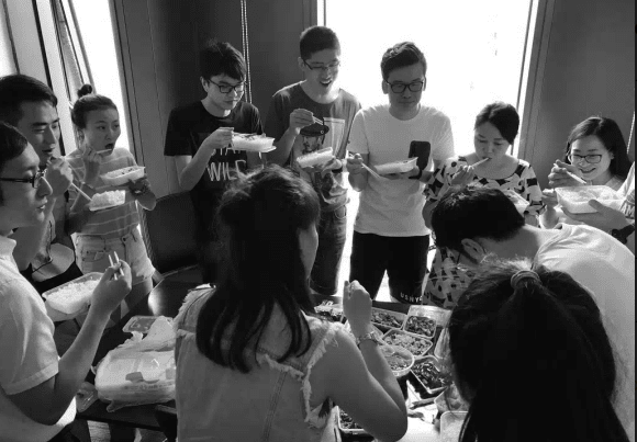 A black and white photograph of a group of young people gathered around a table eating together, with some standing and others seated.