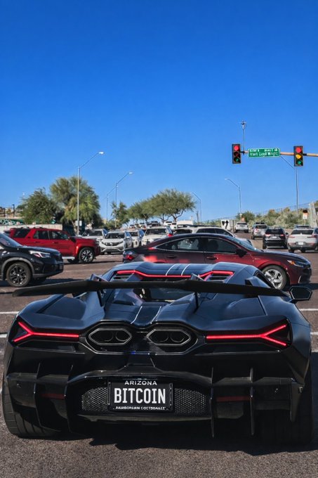 A black Lamborghini waits at a traffic light in Arizona, and its vanity license plate reads "BITCOIN," reinforcing the caption's playful question about whose Lamborghini it is by nodding to crypto wealth culture.
