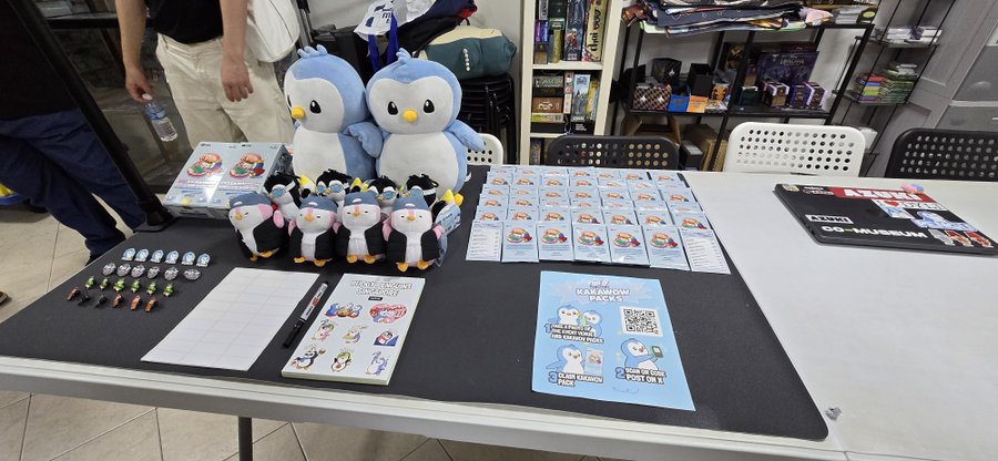 A display table at what appears to be a pop-up shop featuring Kamamon plushies, stickers, and merchandise, with a QR code for a LINE group.