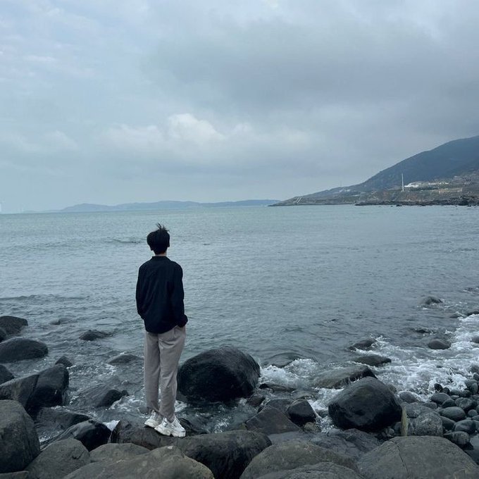 A person in a black jacket and light pants standing on dark rocks at the seashore, looking out at calm waters with mountains in the distant background under a cloudy sky.
