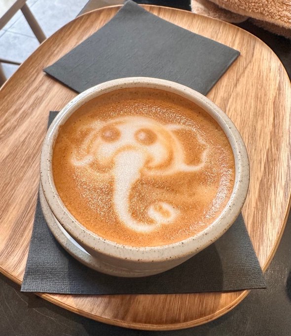 A close-up photo of a ceramic cup of coffee featuring intricate latte art resembling a face with eyes, resting on a gray napkin atop a round wooden tray.