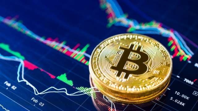 A golden Bitcoin physical coin displayed in front of colorful cryptocurrency price charts on a screen.
