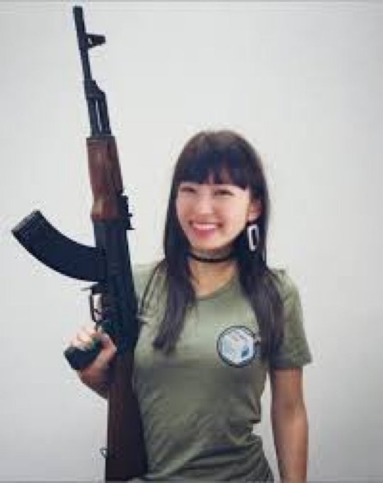 A smiling young woman with long dark hair, wearing an olive green t-shirt and a choker, holds what appears to be an AK-47 style rifle over her shoulder.