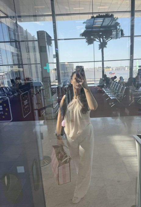 A woman takes a mirror selfie in what appears to be an airport terminal, holding a pink shopping bag with a palm tree visible through the window behind her.