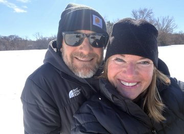 A man wearing a Carhartt beanie and sunglasses and a woman in a black beanie smile together in a snowy outdoor setting, capturing a joyful "Hump Day" moment between the couple.