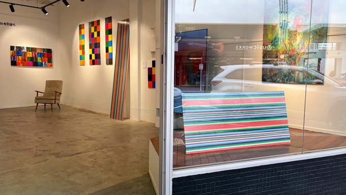 An art gallery interior with colorful geometric and striped paintings on the walls, a single armchair, and a view through the window of a multi-colored striped artwork outside.