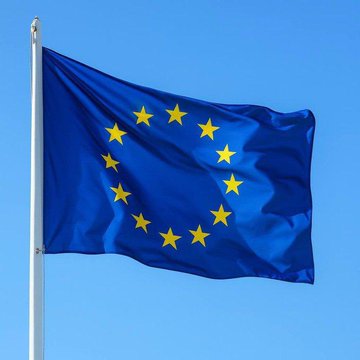 The European Union flag, featuring a circle of 12 yellow stars on a blue background, waving against a clear sky.