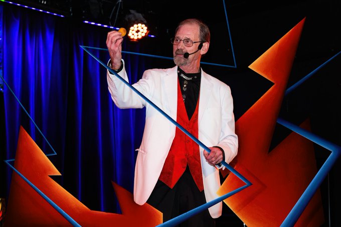 A magician on stage wearing a white blazer over a red vest and headset microphone performs a trick by suspending a ball mid-air with a blue ribbon, fitting the playful collaboration "Mr. Miaside 2" being announced by the performer.