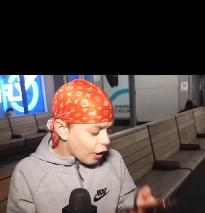 The image features a young boy wearing an orange bandana with a white "Nike" logo, sitting on a bench at Nike Stadium. He appears to be talking into a microphone or recording device, possibly discussing his experience at the stadium or sharing some thoughts about the event. The background of the image shows several benches and chairs arranged in various positions around the area.