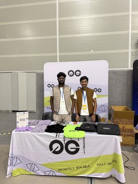 Two young men stand behind a merchandise table displaying t-shirts and accessories, with an OG-branded banner behind them showing 'INFINITELY SCALABLE' and 'FULLY ON'.