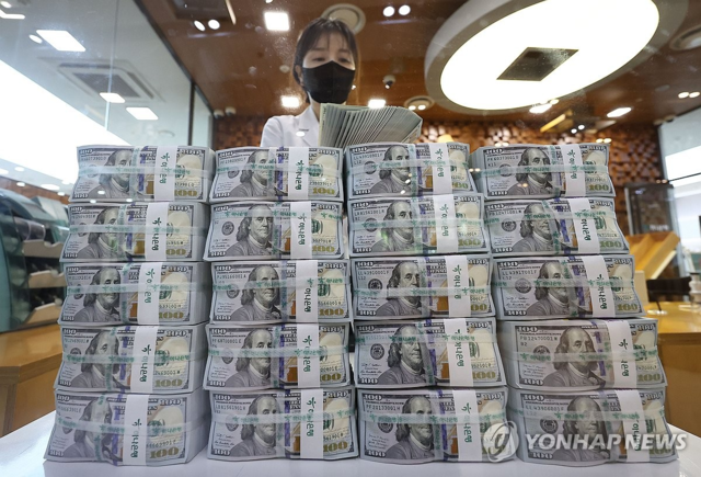 A bank employee wearing a mask holds a fan of US dollar bills behind large stacks of $100 currency on a counter, with a 'Yonhap News' watermark in the corner.