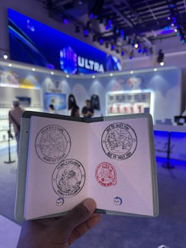 A hand holds open a "Pengu Passport" stamped with official seals in front of an "ULTRA" branded booth, illustrating the in-person stamping opportunities for Pudgy Penguins and PenguAsia holders at official events.